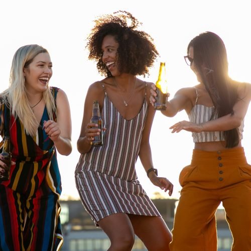 Group of cheerful young multiracial ladies laughing and dancing with beer bottles in hands during summer party on rooftop on sunny day